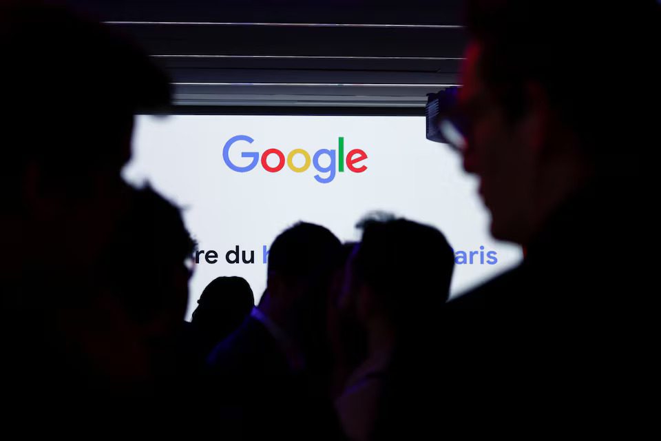 Silhouettes of people gathered in front of a brightly lit screen displaying the multicolored Google logo and some faint text below it, possibly at a conference or event.