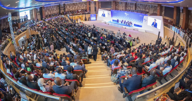 A wide, high-angle view of a vast, tiered conference hall packed with a large audience of hundreds of people in business attire. The audience is facing a central stage where a panel discussion is taking place. Large screens and signage with an 'AFRICA' theme are visible on the stage backdrop.