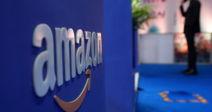 A shallow depth-of-field close-up of the raised white Amazon logo and orange smile arrow mounted on a vibrant blue surface. A blurry figure of a person in a dark suit stands on a blue carpet in the background.