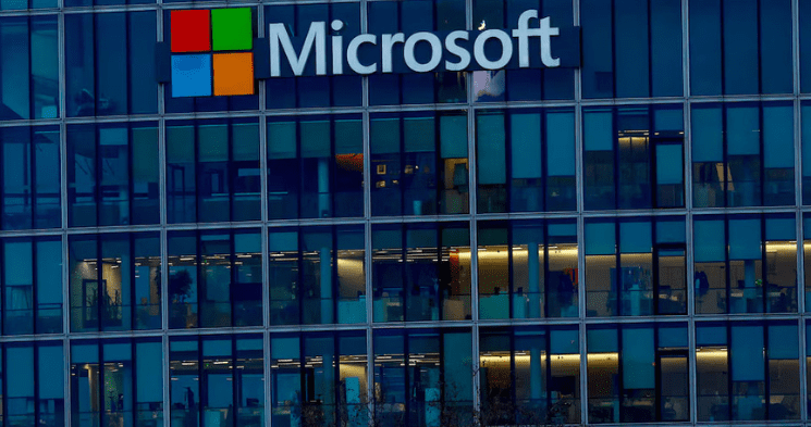 A close-up view of the exterior of a glass office building at dusk or night, prominently featuring the Microsoft logo and name on the top.