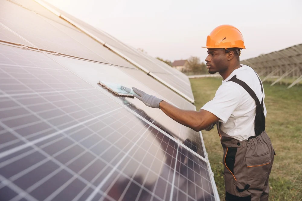 A male maintenance worker in an orange hard hat and gray overalls cleans a large solar panel with a soft cloth at a solar farm during sunset.