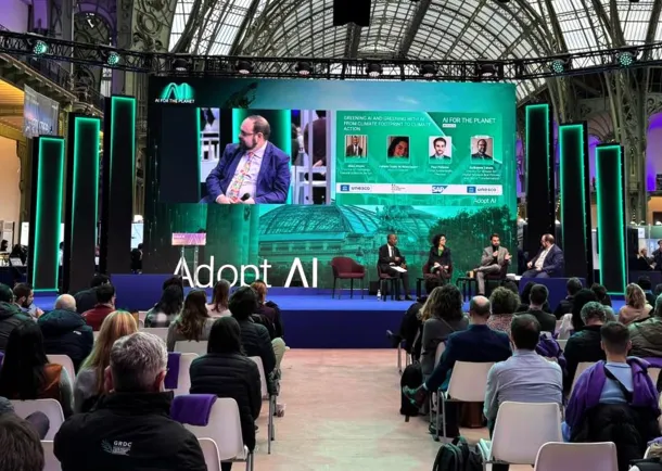 An indoor conference stage at the "Adopt AI" event featuring a panel discussion titled "Greening AI and Greening with AI." Four panelists sit on a blue stage in front of a large digital screen displaying their profiles and the "AI for the Planet" logo. An audience is seated in the foreground, facing the stage within a grand, high-ceilinged architectural space.