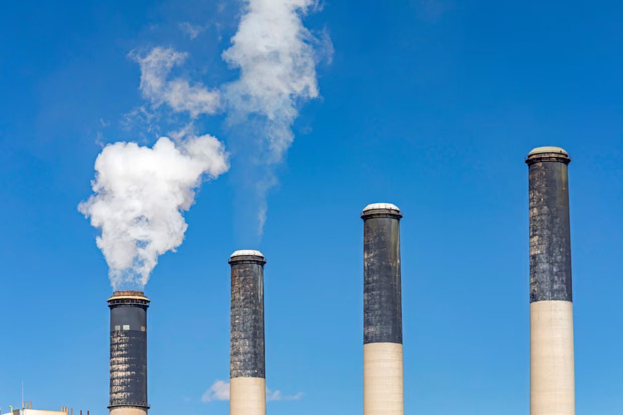 Four industrial smokestacks stand against a clear blue sky, with white smoke or steam billowing from the leftmost two.